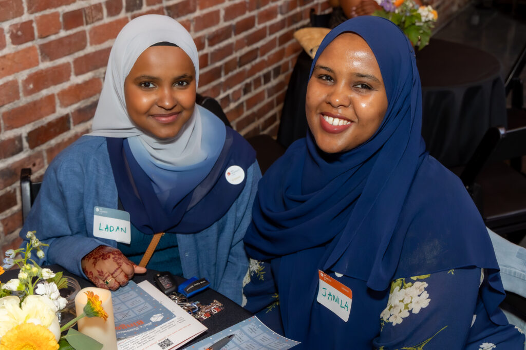 Two smiling women, dressed in blue, sitting at a table in front of a brick wall.