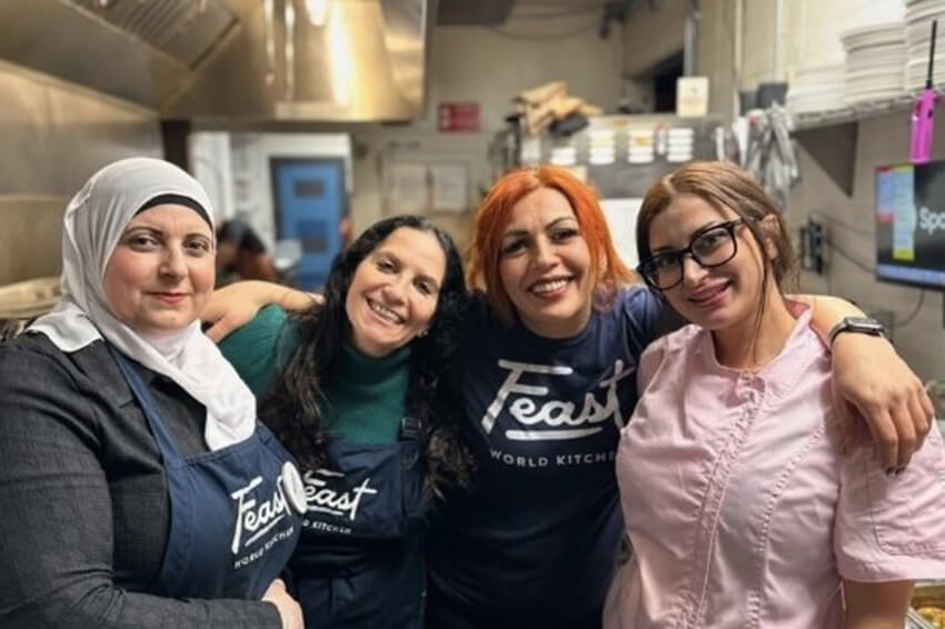 Four women standing in an industrial kitchen with their arms around each other and smiling at the camera.
