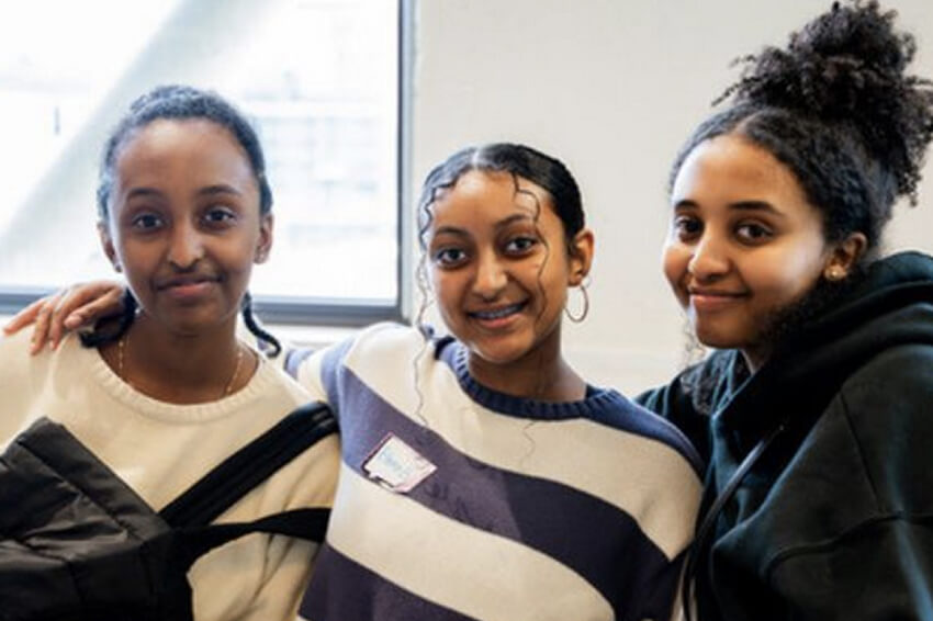 Three young women standing in front of a white wall with their arms around each other and smiling at the camera.