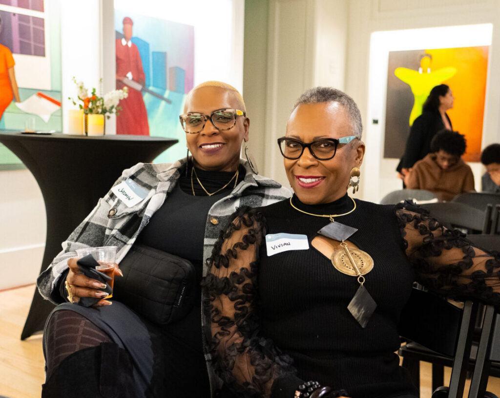 Two women sitting on black chairs in an art gallery
