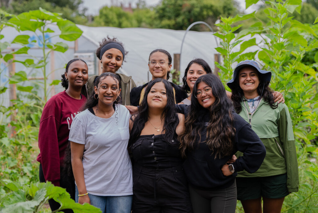 A group of 8 women standing in a garden with their arms around each other and smiling at the camera.
